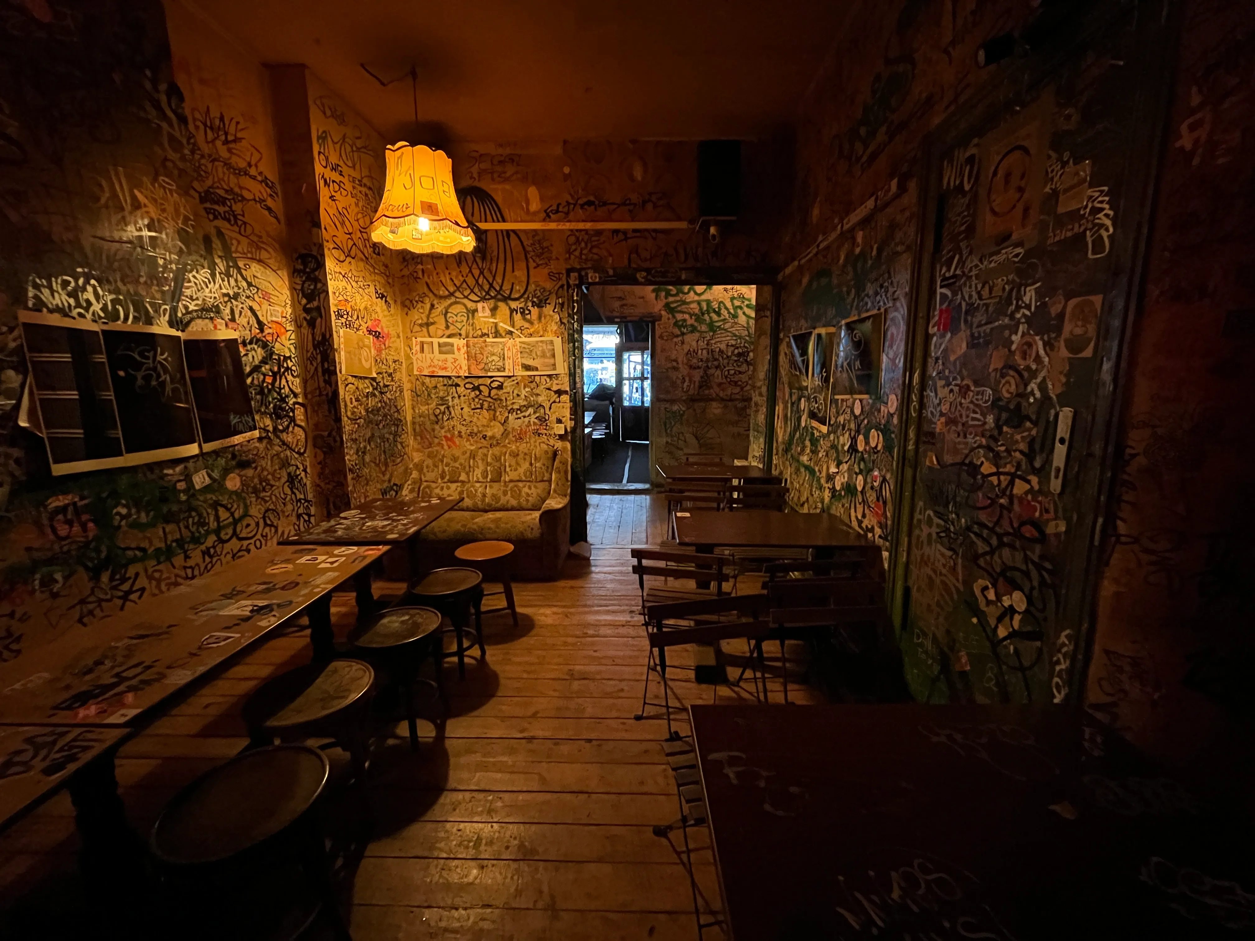 Bernd's Wohnzimmer: warm beleuchteter Berliner Eventraum mit graffitibedeckten Wänden, Holzdielen, Bistrotischen und einer alten Stehlampe mit Stoffschirm.