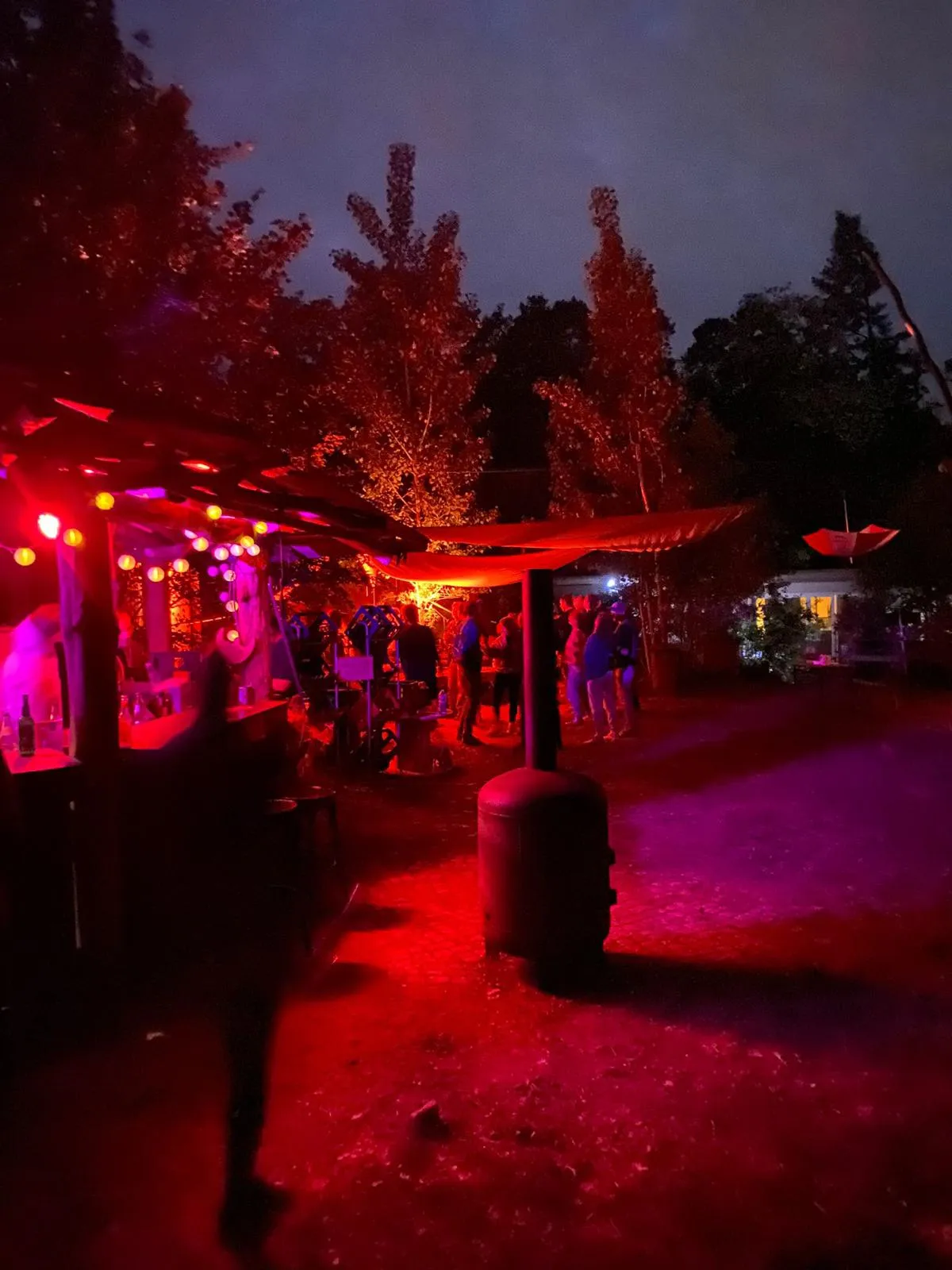 Försterei bei Nacht: Outdoor-Bar mit roter Beleuchtung, Zeltdach mit Lichterketten und Gäste zwischen hohen Bäumen.